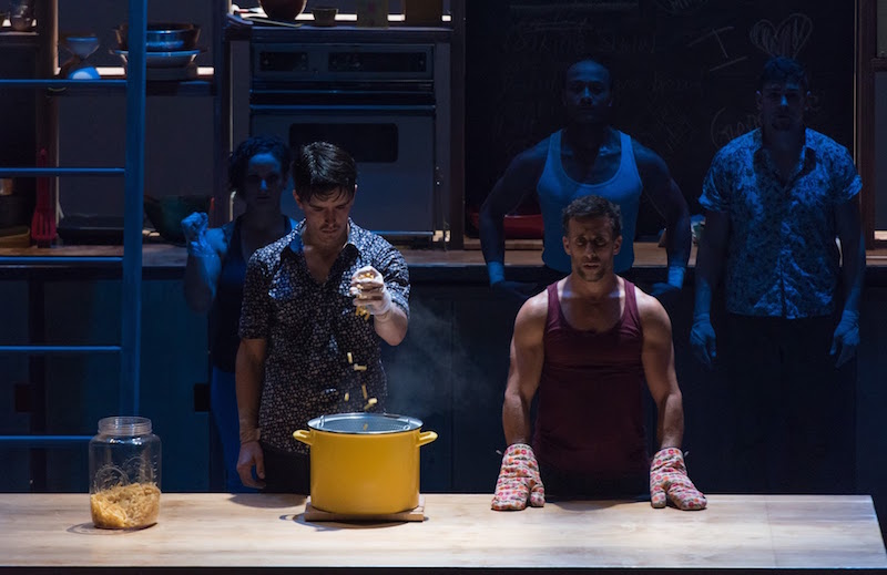 A group of performers watch a man throw pasta in a yellow pot